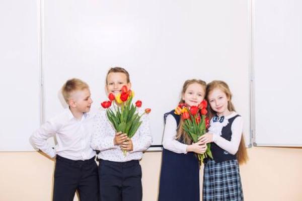 School children hold flowers and stand together in a classroom.